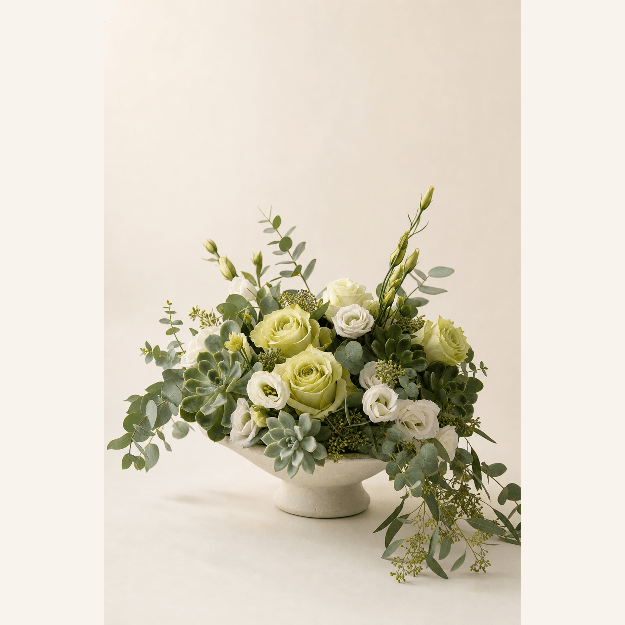 Soft green and white roses, lisianthus, succulents and eucalyptus in a cream ceramic footed bowl