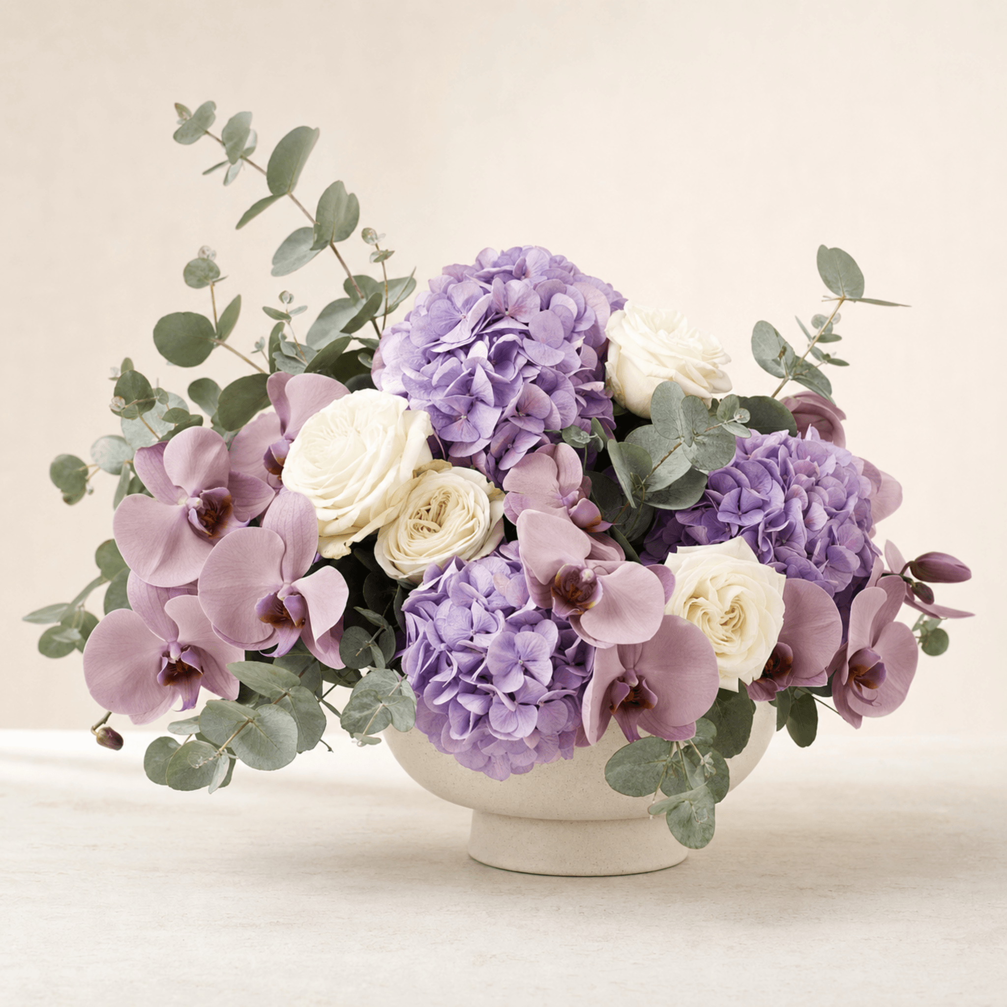 Purple hydrangeas, mauve phalaenopsis orchids, cream roses and eucalyptus in a cream ceramic bowl